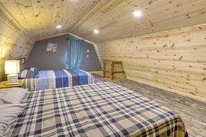 A bedroom with a sloped ceiling, a bed with a plaid bedspread, a nightstand with a lamp, and a window with curtains.