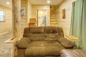 A cozy log cabin interior with a large cushioned sofa, wooden walls, a small table, and a green curtain.