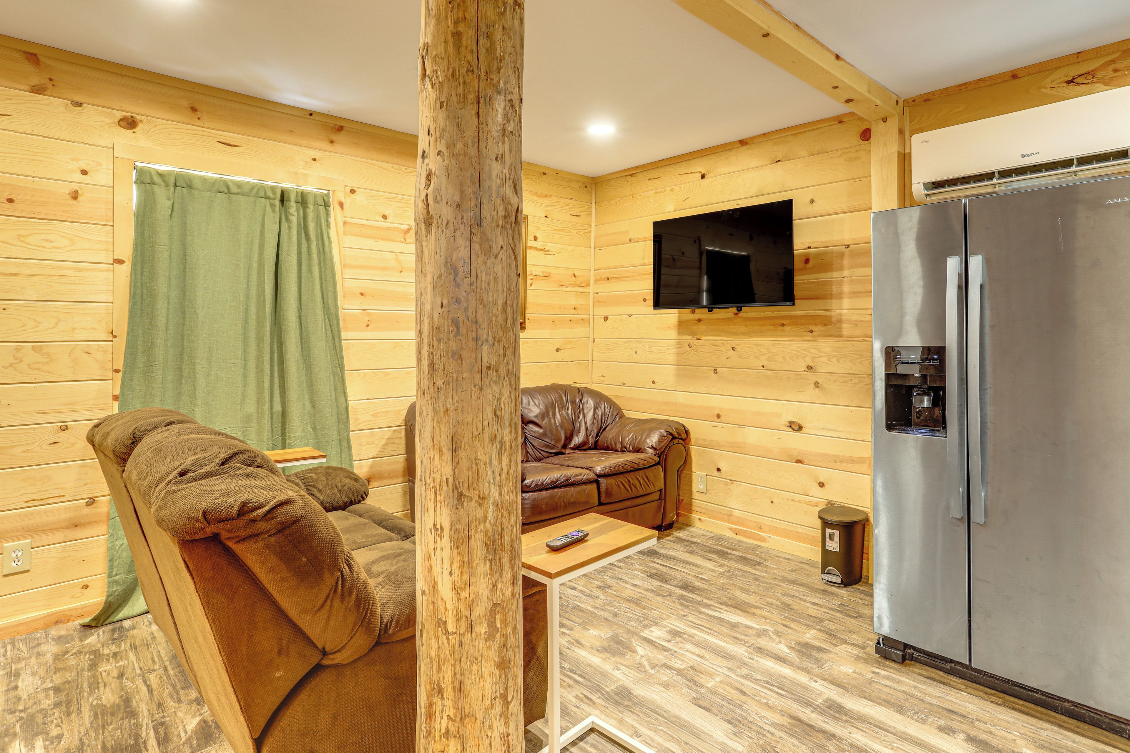 A cozy room with a brown leather sofa, a small wooden table, a television mounted on the wall, and a stainless steel refrigerator.
