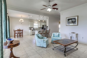 A spacious living room with a ceiling fan, a dining area with chairs, a kitchen in the background, and a framed picture on the wall.