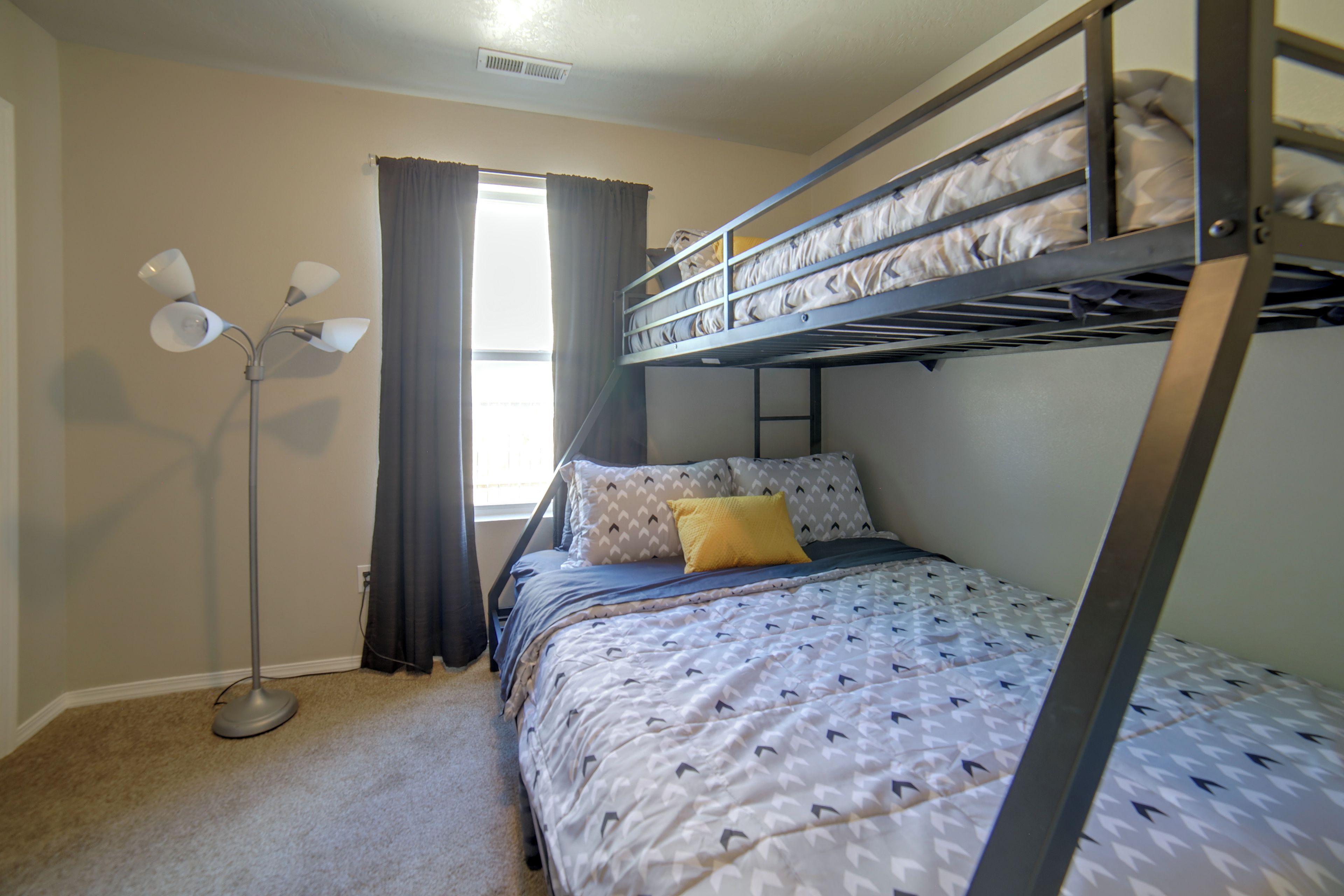 A bunk bed with a patterned bedspread and pillows, a standing floor lamp, and a window with curtains.