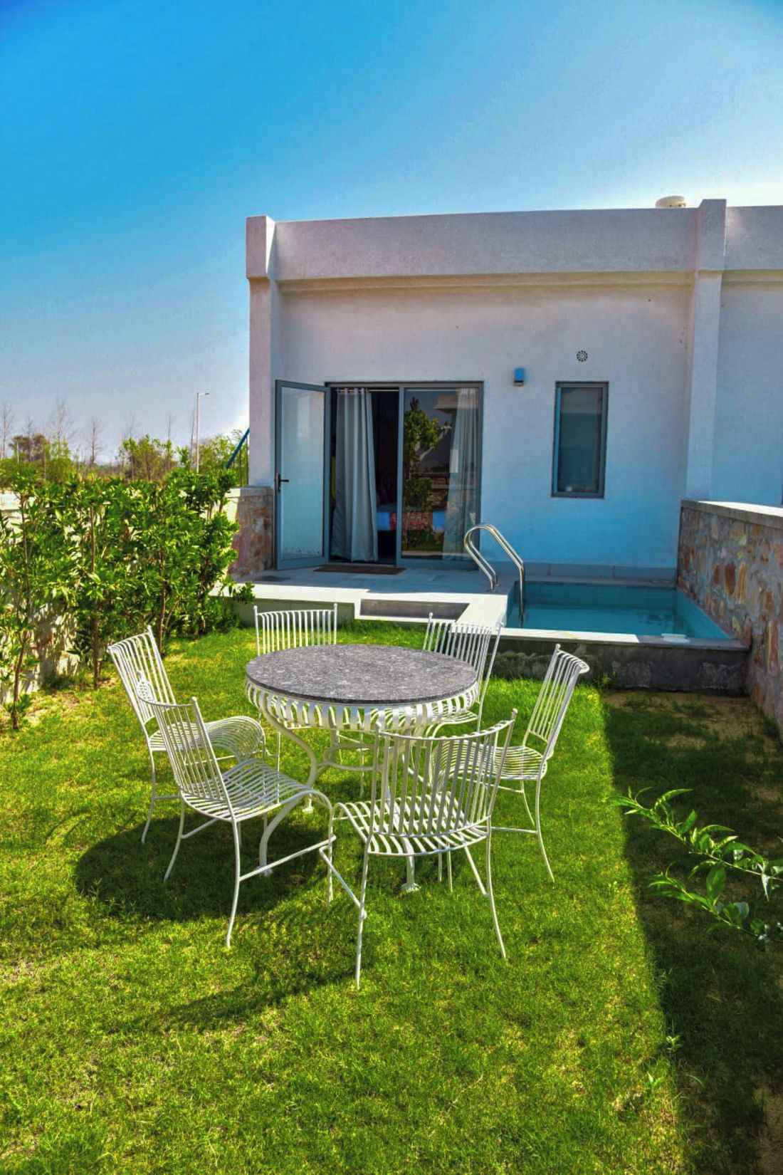 A modern outdoor patio with a round table and metal chairs, a swimming pool, and a building with a sliding glass door.