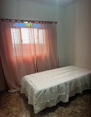 A room with a bed, a window with sheer curtains, and a tiled floor.