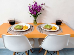 A dining table with two plates of food, a vase with flowers, and two cups of coffee. - Easy Playa Mornings. 2 Beds Sleeps 4 · 5th Ave, ADO, Beach, Cozumel Ferry 4 Mins (Playa del Carmen)