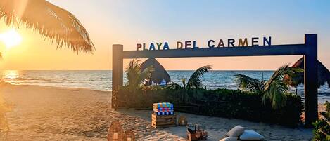 A beach entrance with a sign reading 'Playas del Carmen,' a beach umbrella, lounge chairs, and a wicker basket with a bottle.