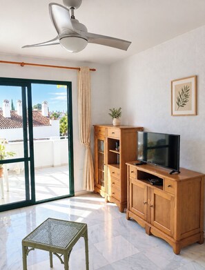 Een kamer met een plafondventilator, een televisie op een houten kast, een tafeltje en een glazen schuifdeur naar een buitenruimte.