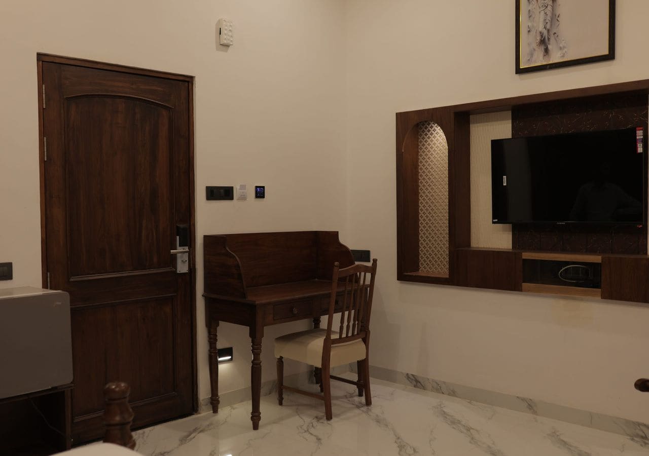A room with a wooden door, a desk with a chair, a television mounted on the wall, and a marble floor.