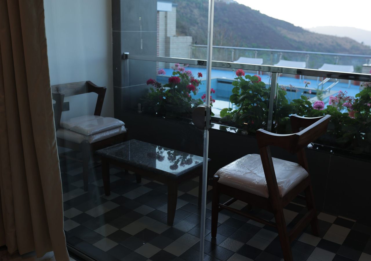 A balcony with a checkered floor, two wooden chairs, a glass table, and potted plants with pink flowers.