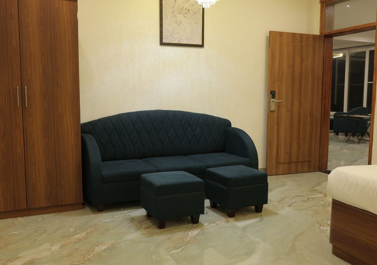 A room with a dark-colored sofa, two matching ottomans, a wooden wardrobe, and a door leading to another room.