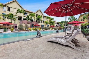 A pool area with lounge chairs and umbrellas, surrounded by buildings with balconies and palm trees. - Cosy 1 Bedroom Unit centrally located in Cairns. (Cairns North)