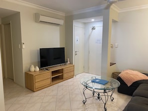 A living room with a flat-screen TV on a wooden stand, a glass coffee table with a book, a black sofa, and a shower area with a door. - Cosy 1 Bedroom Unit centrally located in Cairns. (Cairns North)