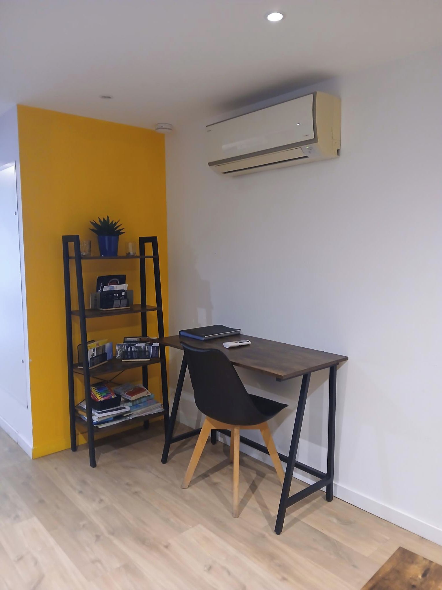 Una habitación con un escritorio, una silla y una estantería frente a una pared amarilla.