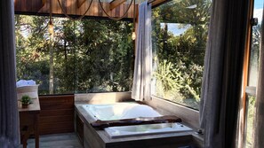 En træhytte med et stort vindue, en træbænk, et lille bord med en plante og en jacuzzi.