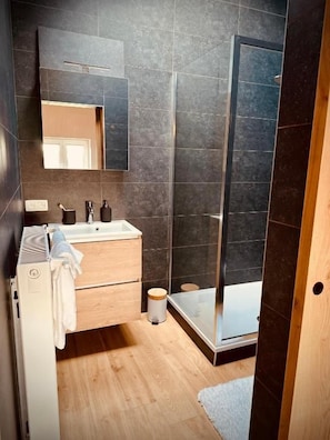 A modern bathroom with a wooden vanity, a large mirror, a shower enclosure, and a tiled floor. - Prim'tout (Ohey)