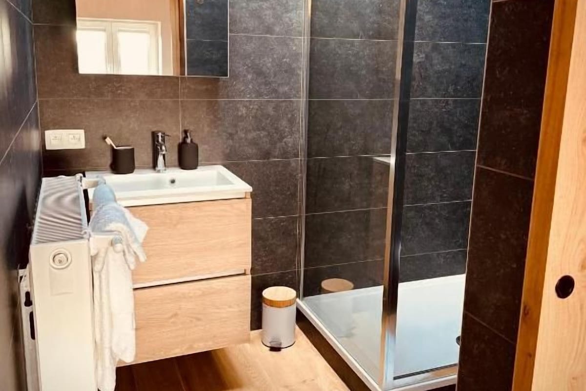 A modern bathroom with a wooden vanity, a large mirror, a shower enclosure, and a tiled floor.