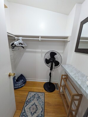 A small room with a wooden floor, a black standing fan, a white wall, a wooden stool, and a blue patterned rug. - Room #3 - Queen Size Bedroom in Shared Townhouse (Toronto)