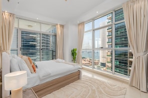 A modern bedroom with a large bed, a bedside lamp, and a view of a cityscape through floor-to-ceiling windows. - Premium 4BR at Sky View Tower (Dubai)