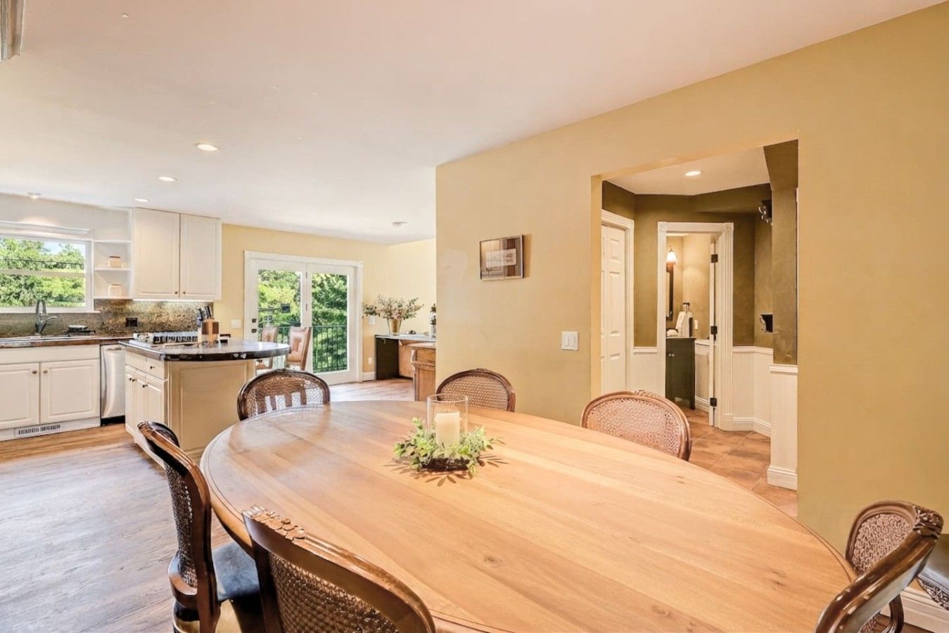 A spacious dining area with a wooden table and chairs, a kitchen with white cabinets, and a view of greenery outside.