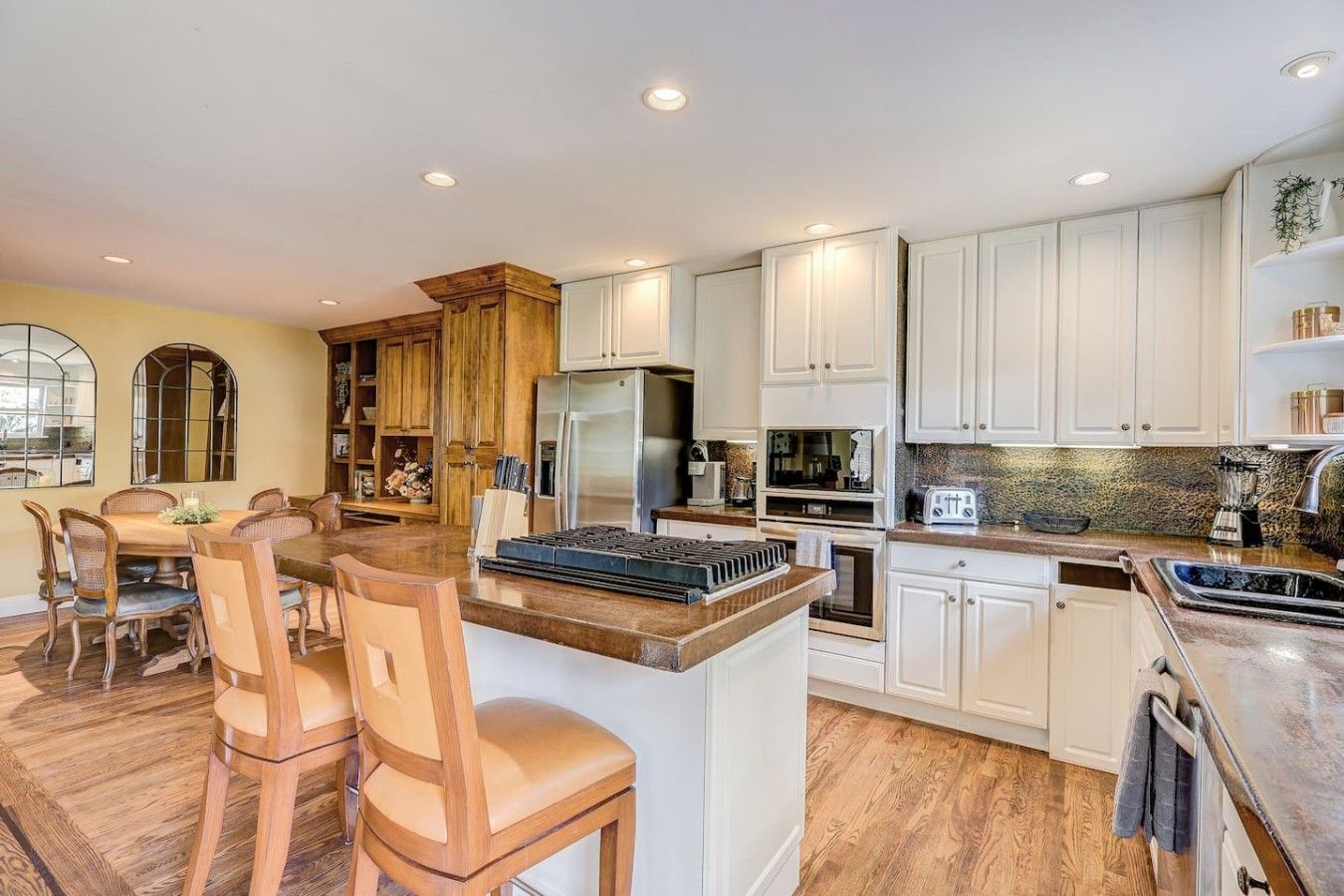 A modern kitchen with white cabinets, a central island with a stove, and a dining area with wooden chairs.