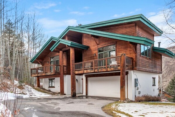 A two-story wooden cabin with a balcony, a garage, and a snow-covered driveway.