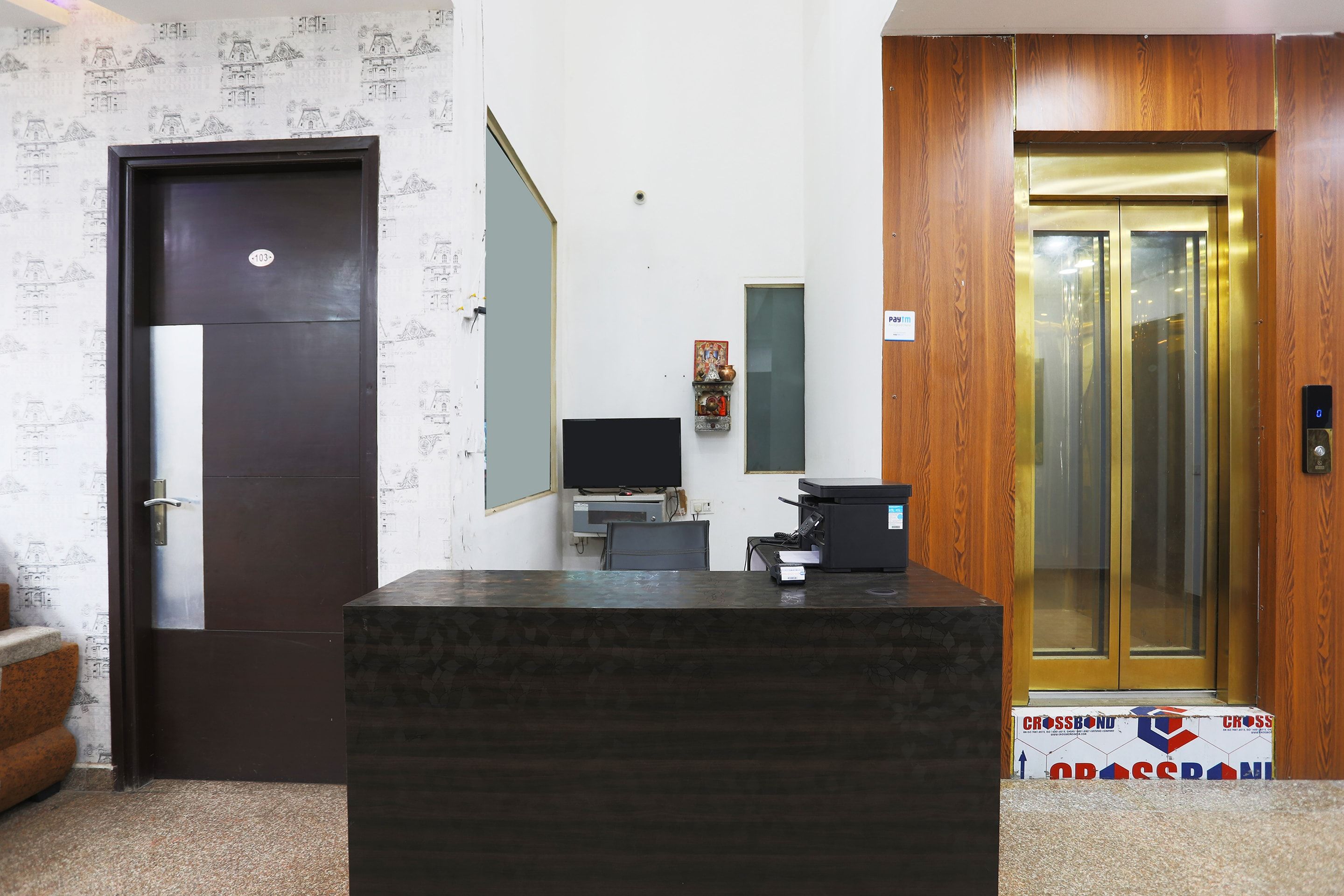 A reception desk with a computer, a telephone, and a small shelf with decorative items. There is a door with a number plate and a wooden panel wall with a patterned wallpaper.