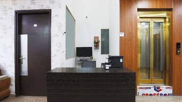 A reception desk with a computer, a telephone, and a small shelf with decorative items. There is a door with a number plate and a wooden panel wall with a patterned wallpaper.