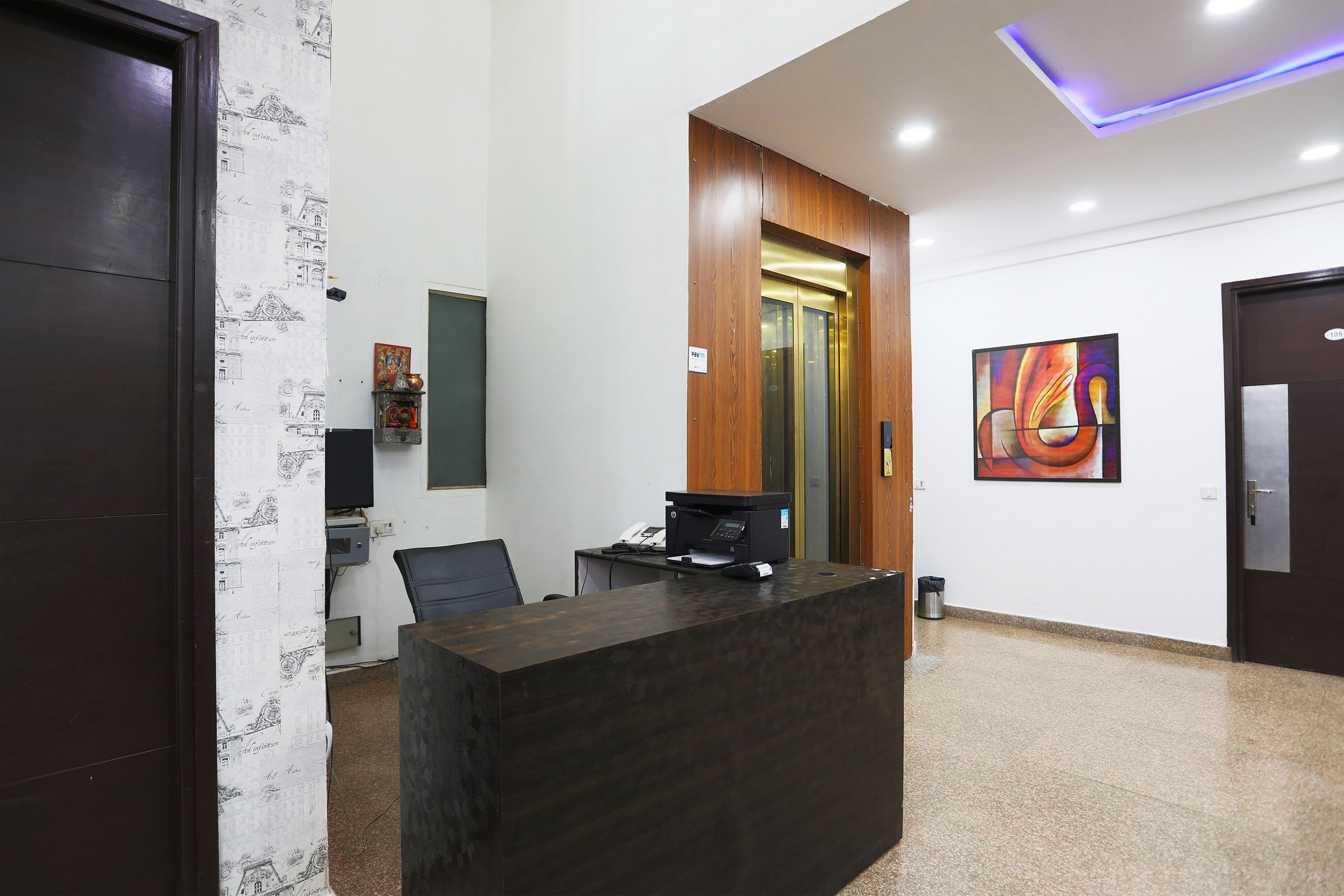 A reception desk with a computer, a telephone, and a chair in a modern office space with a wooden door and a painting on the wall.