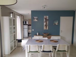 A dining area with a white table and chairs, a kitchen in the background, and a wall with directional signs. - Air-conditioned chalet for 6–8 people in a vacation resort (Parentis-en-Born)