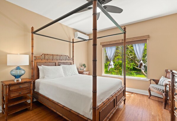 Uma cama de dossel com cabeceira, mesa de cabeceira com luminária, uma cadeira e uma janela com cortinas.