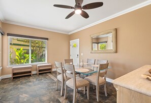 A dining area with a ceiling fan, a dining table with chairs, a bench, and a window with blinds. - Tropical Oasis at Regency 514 - Steps from Poipu Beach (Koloa)