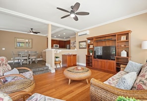A living room with a television, wicker furniture, a ceiling fan, and a kitchen in the background. - Tropical Oasis at Regency 514 - Steps from Poipu Beach (Koloa)