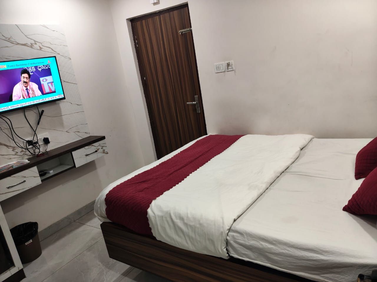 Ein Schlafzimmer mit einem Bett, einem Fernseher an der Wand, einer Holztür und einem kleinen Schreibtisch mit Schubladen.