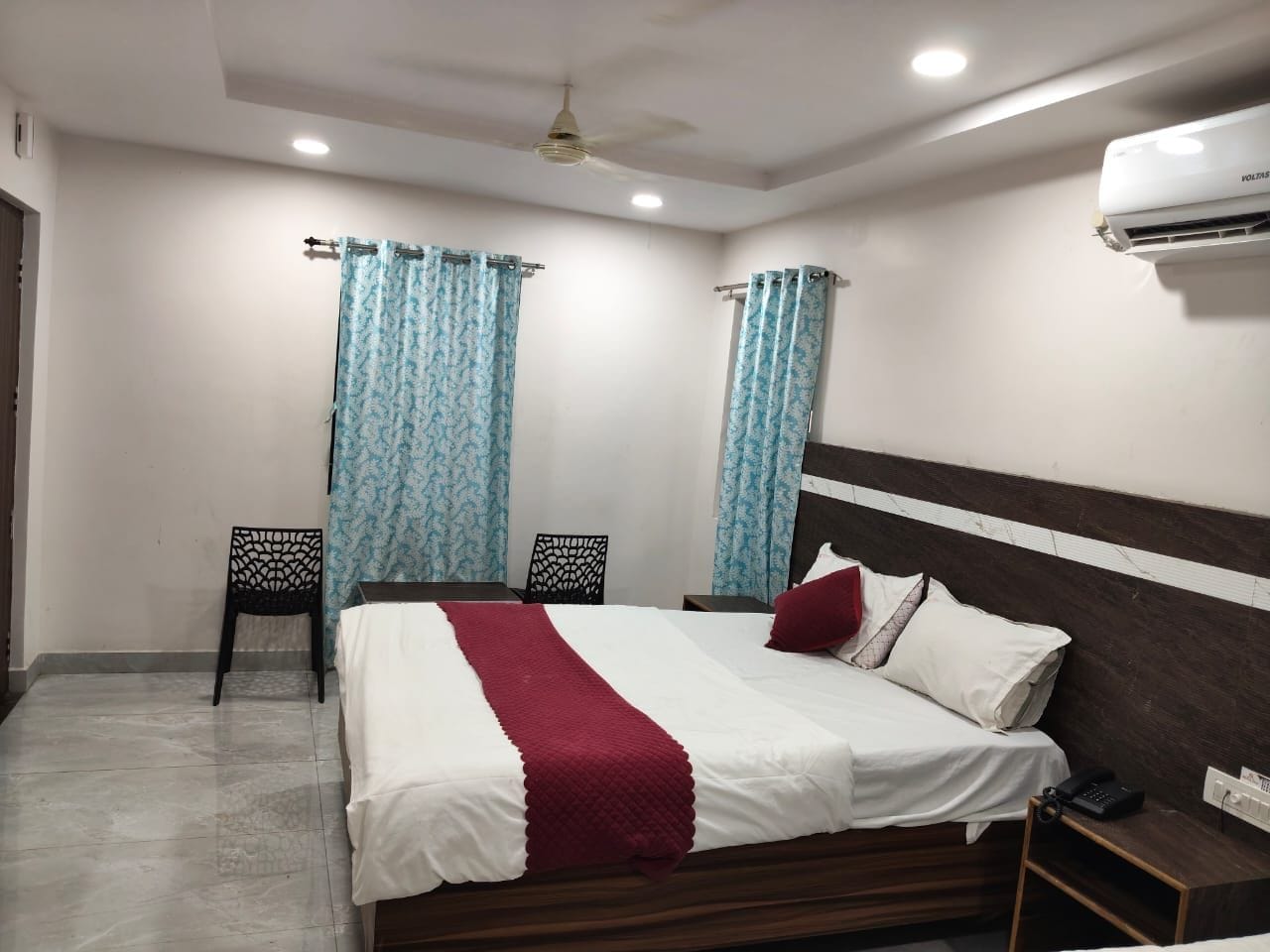 Una habitación de hotel con cama, dos sillas, mesita de noche, aire acondicionado y cortinas.