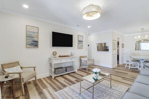 Una sala de estar con televisor de pantalla plana, un comedor con candelabro y una cocina visible a través de un vano abierto.