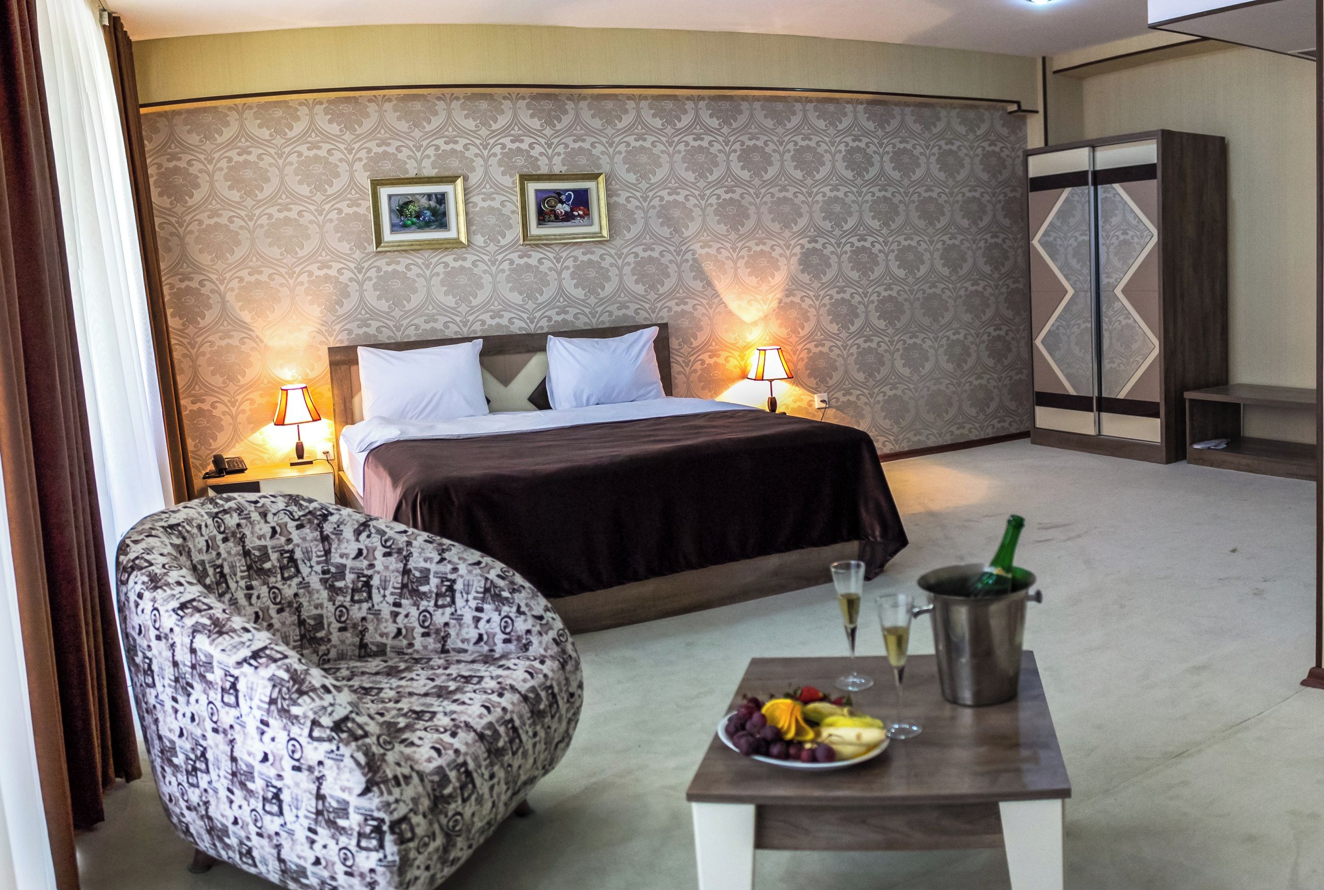 Une chambre d’hôtel comprenant un lit, deux lampes de chevet, un fauteuil à motifs, une petite table avec des fruits et des boissons, et une armoire.
