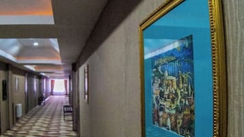 A hotel hallway with a patterned carpet, a framed artwork on the wall, and a bench.