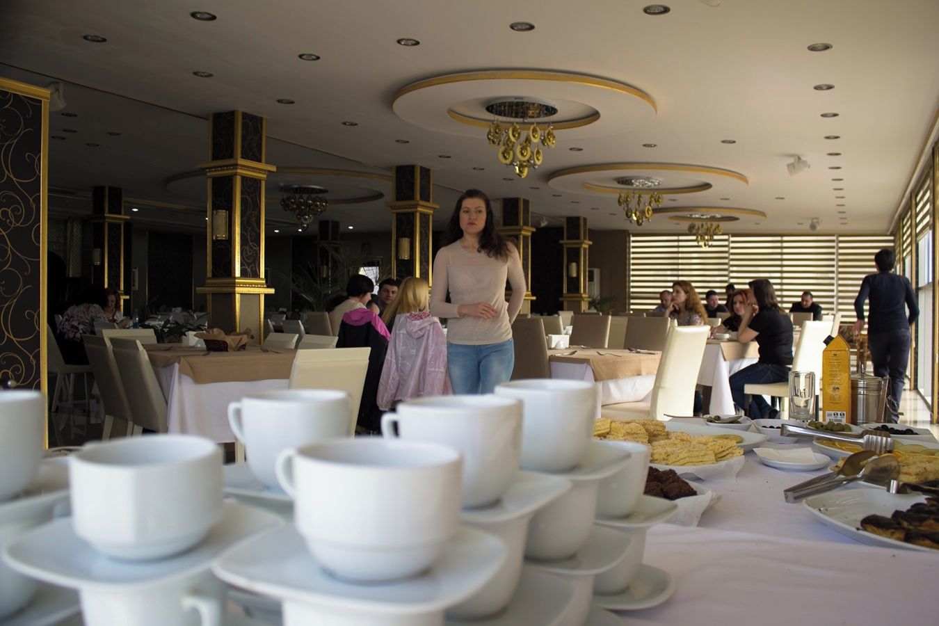 Un comedor con mesas dispuestas para una comida, una mujer de pie al frente y una araña colgante.
