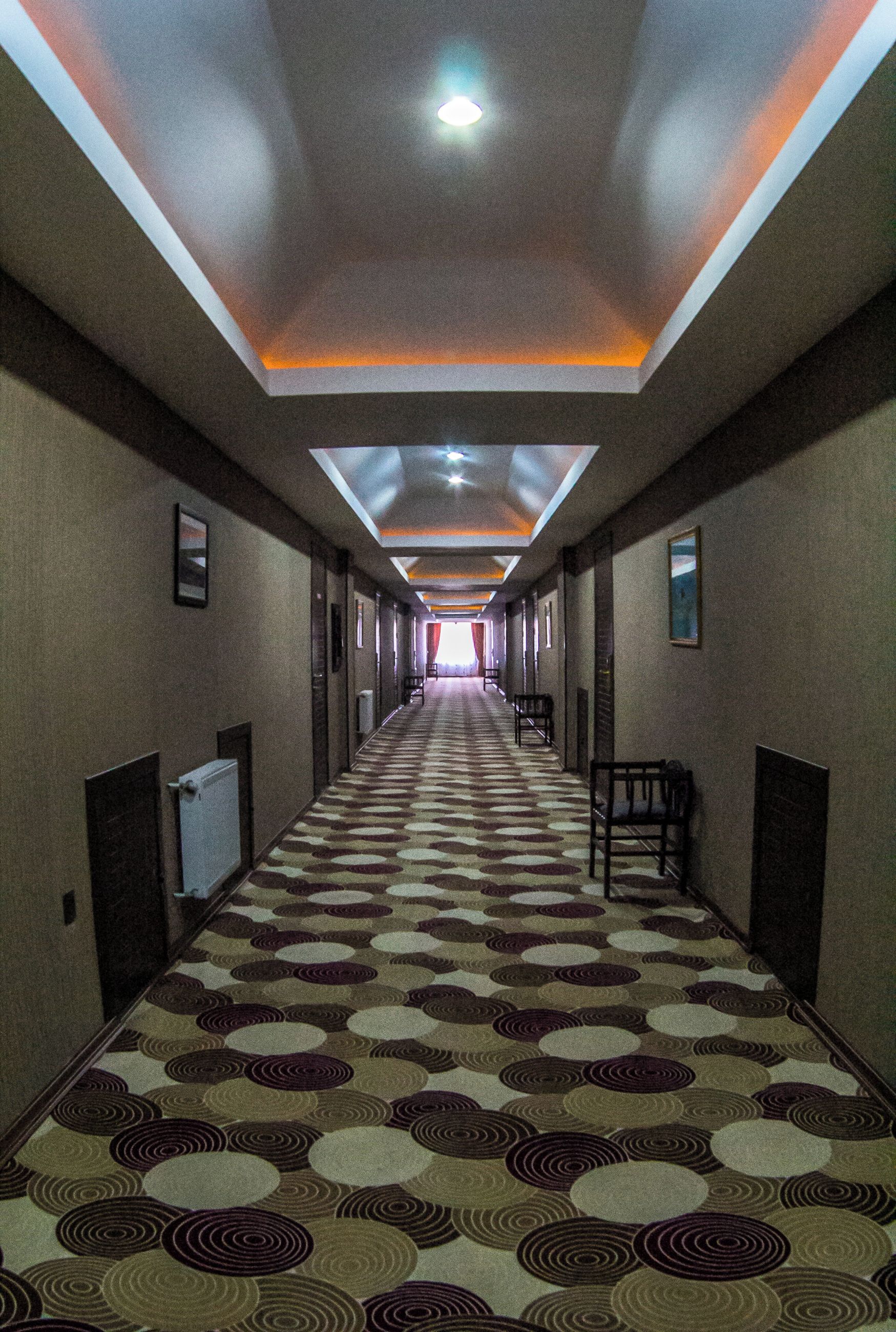 Un pasillo de hotel con alfombra estampada, iluminación empotrada y sillas a lo largo de las paredes.