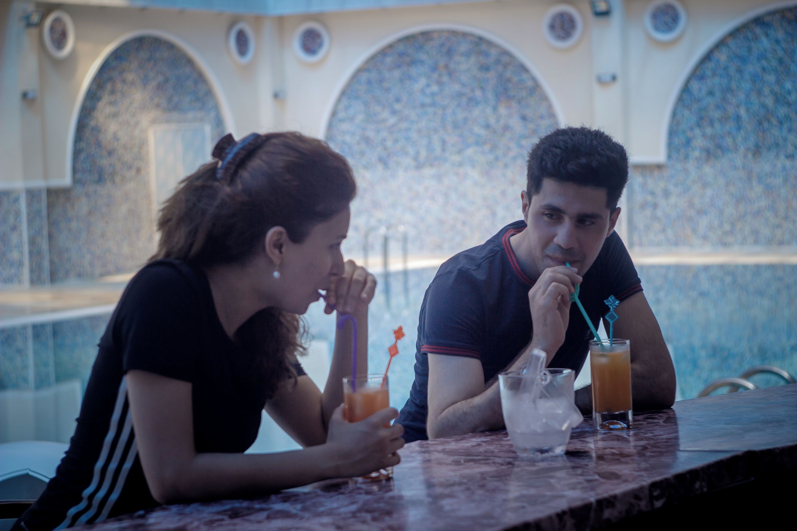Dos personas sentadas en una mesa con bebidas, con vista a una zona de piscina con ventanales arqueados.
