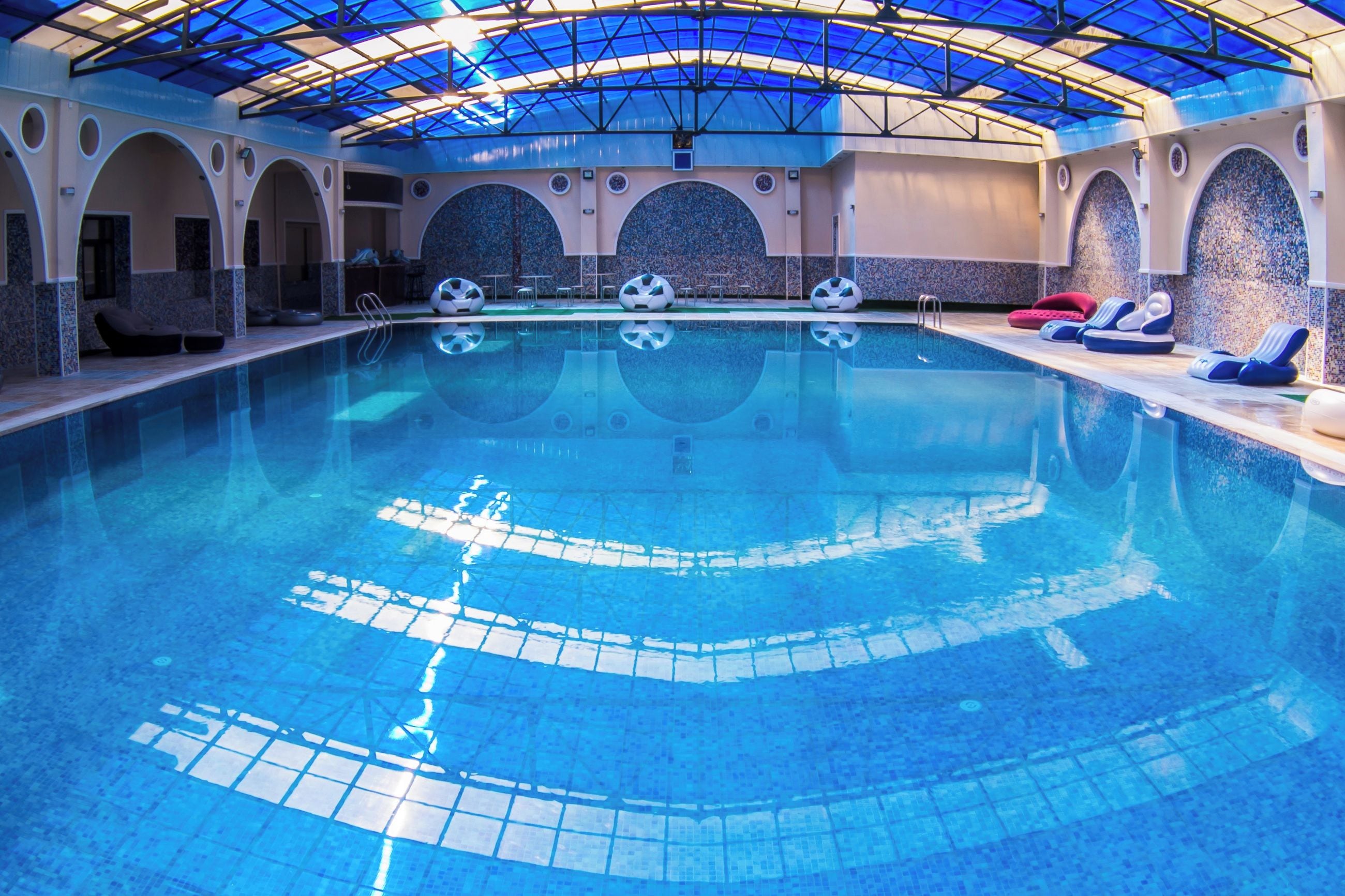 Une piscine intérieure à l’eau d’un bleu cristallin, dotée d’un toit en verre et d’espaces aménagés avec des chaises longues.