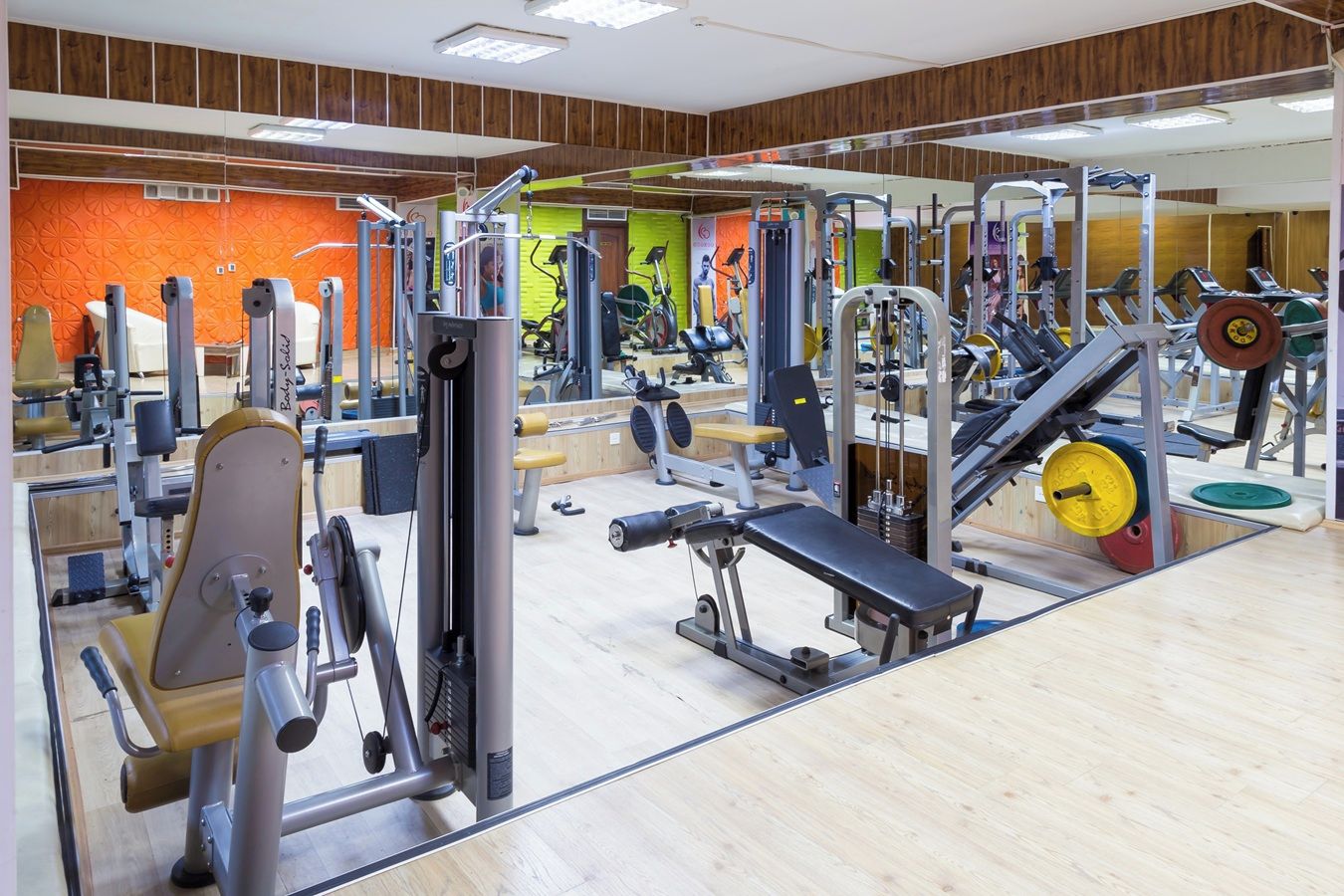 A gym with various exercise equipment, including weight machines and free weights, and a mirrored wall.