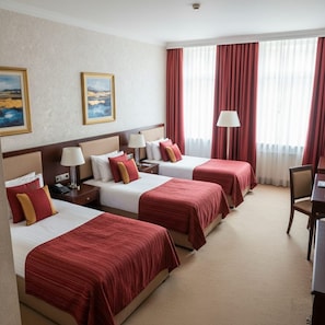 Habitación de hotel con tres camas, cada una con ropa de cama roja, un escritorio y una silla. Hay dos cuadros en la pared y una ventana con cortinas.