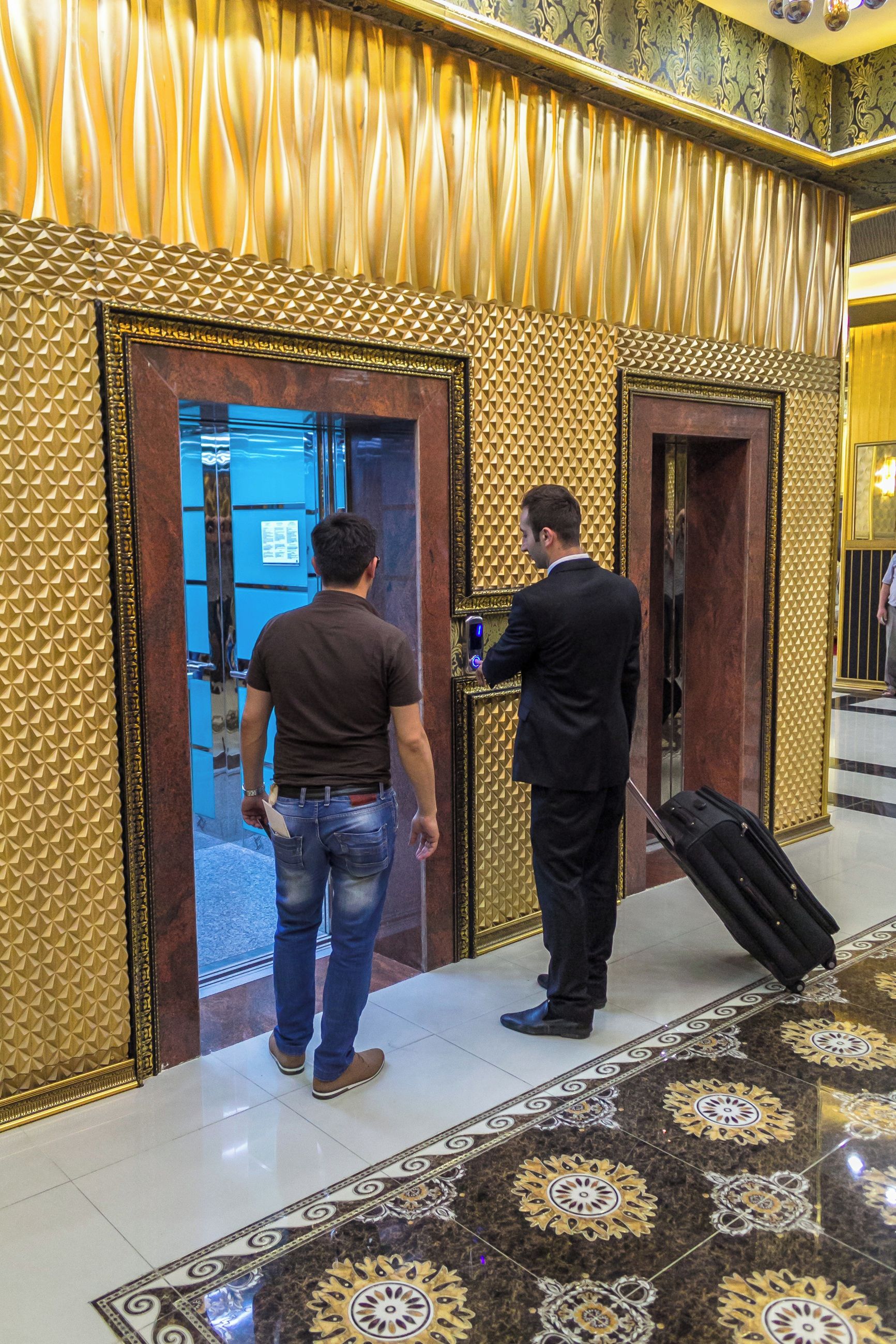 Un hombre con traje y ropa informal de pie frente a la entrada de un ascensor en el vestíbulo de un hotel, con suelo estampado y diseño dorado en la pared.