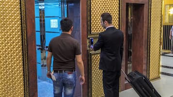 Un hombre con traje y ropa informal de pie frente a la entrada de un ascensor en el vestíbulo de un hotel, con suelo estampado y diseño dorado en la pared.