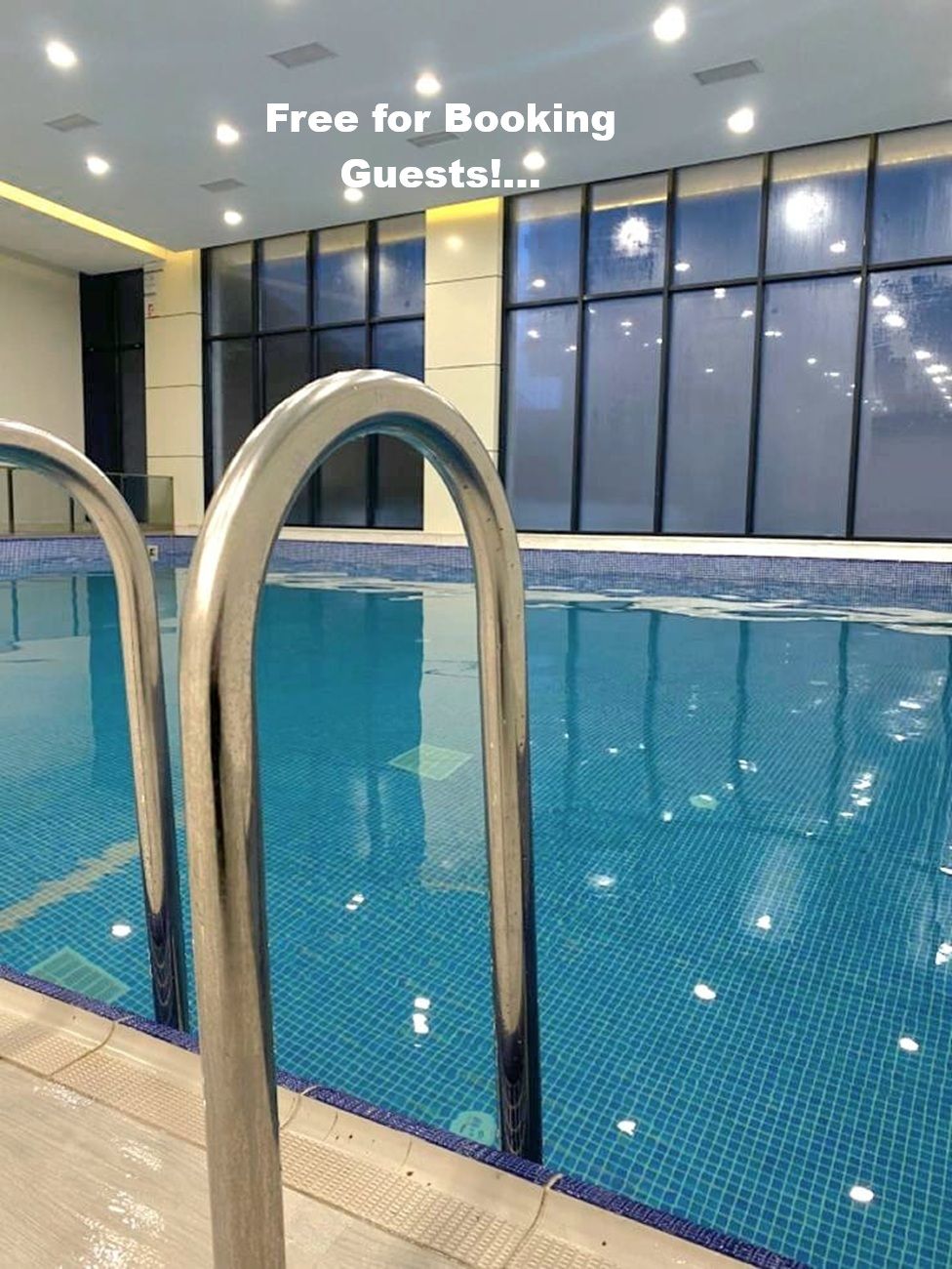 An indoor swimming pool with clear water, metal pool ladders, and a tiled wall with windows in the background.