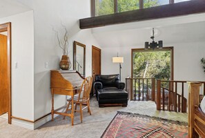Um quarto amplo com um piano, uma poltrona de couro preto, um tapete estampado e vista para árvores através de uma grande janela.