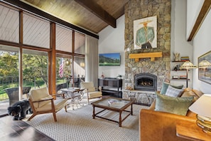 A spacious living room with a stone fireplace, comfortable seating, and a large window overlooking a garden. - New! FOREteener’s Retreat | AC, Pool, Hot Tub, Golf & Ski (Durango)