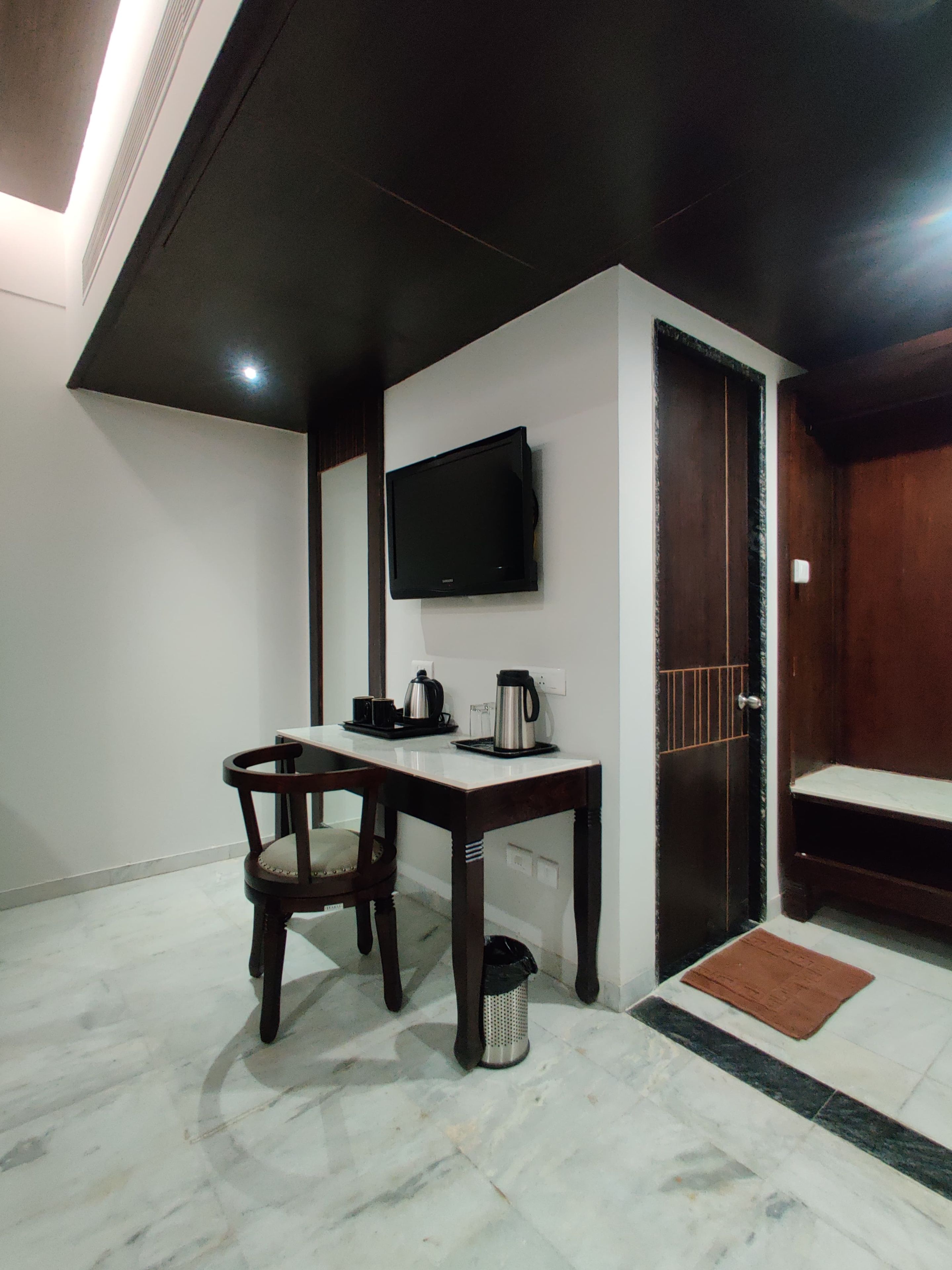 A room with a flat-screen TV mounted on the wall, a wooden table with a chair, a coffee maker, and a trash bin.