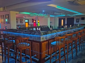 Una zona de bar con una barra larga, varios taburetes y una variedad de botellas y vasos en los estantes.