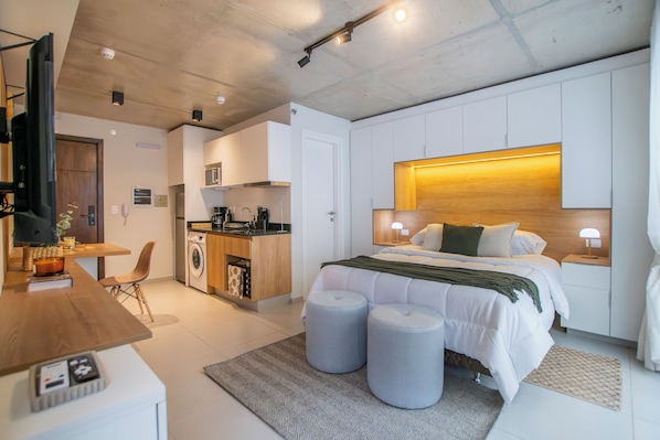 A modern hotel room with a bed, bedside lamps, a small kitchen area, and a seating area with a table and chair. - Corporate Stays First del Sol - 604 (Asunción)
